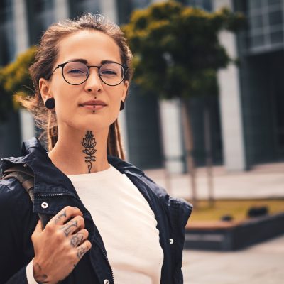 Nice young woman with piercing, glasses and tattoo on her neck is posing for photographer on the street.